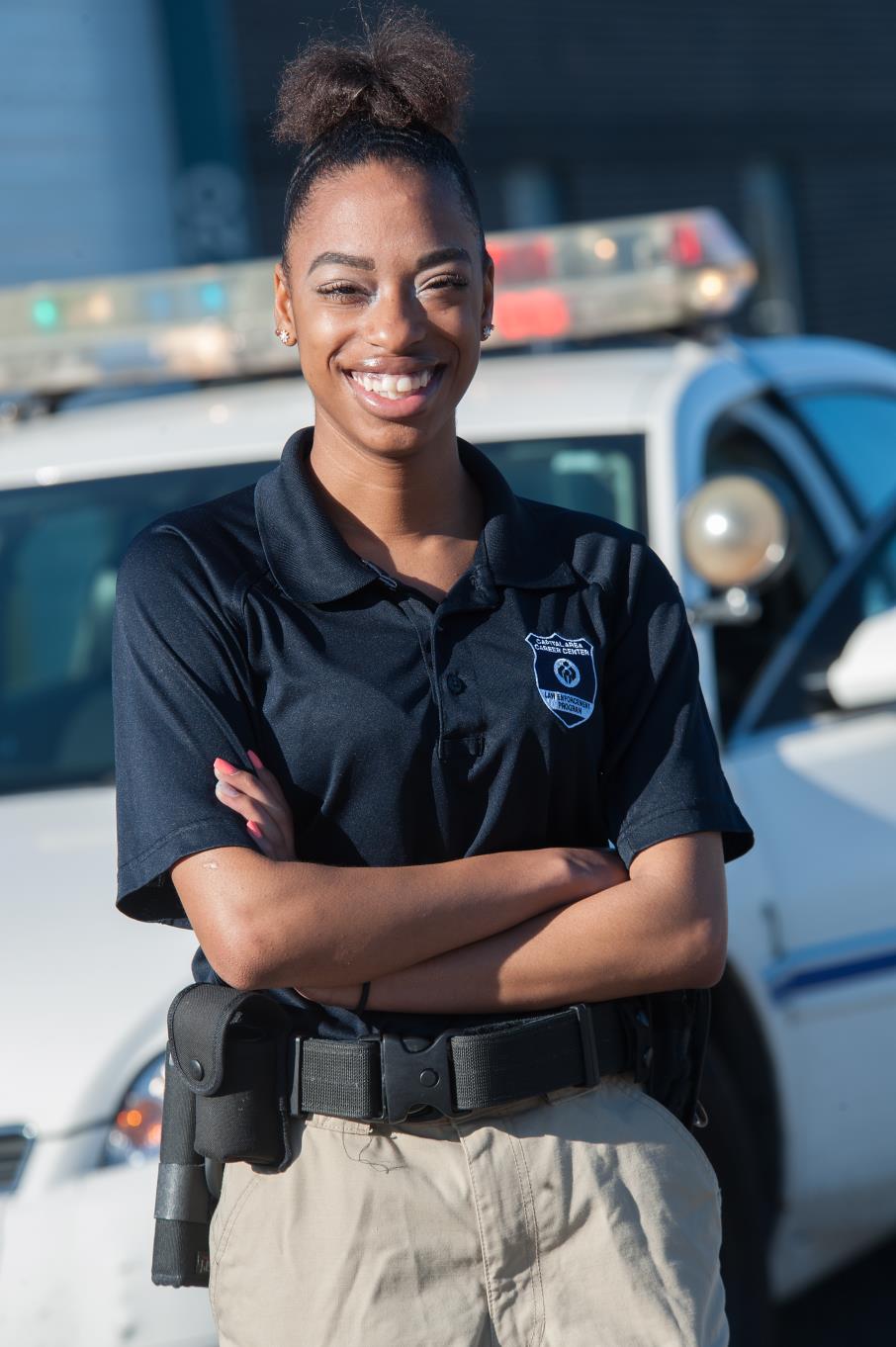 Female Law Enforcement student stands in front of her patrol car with her arms crossed.