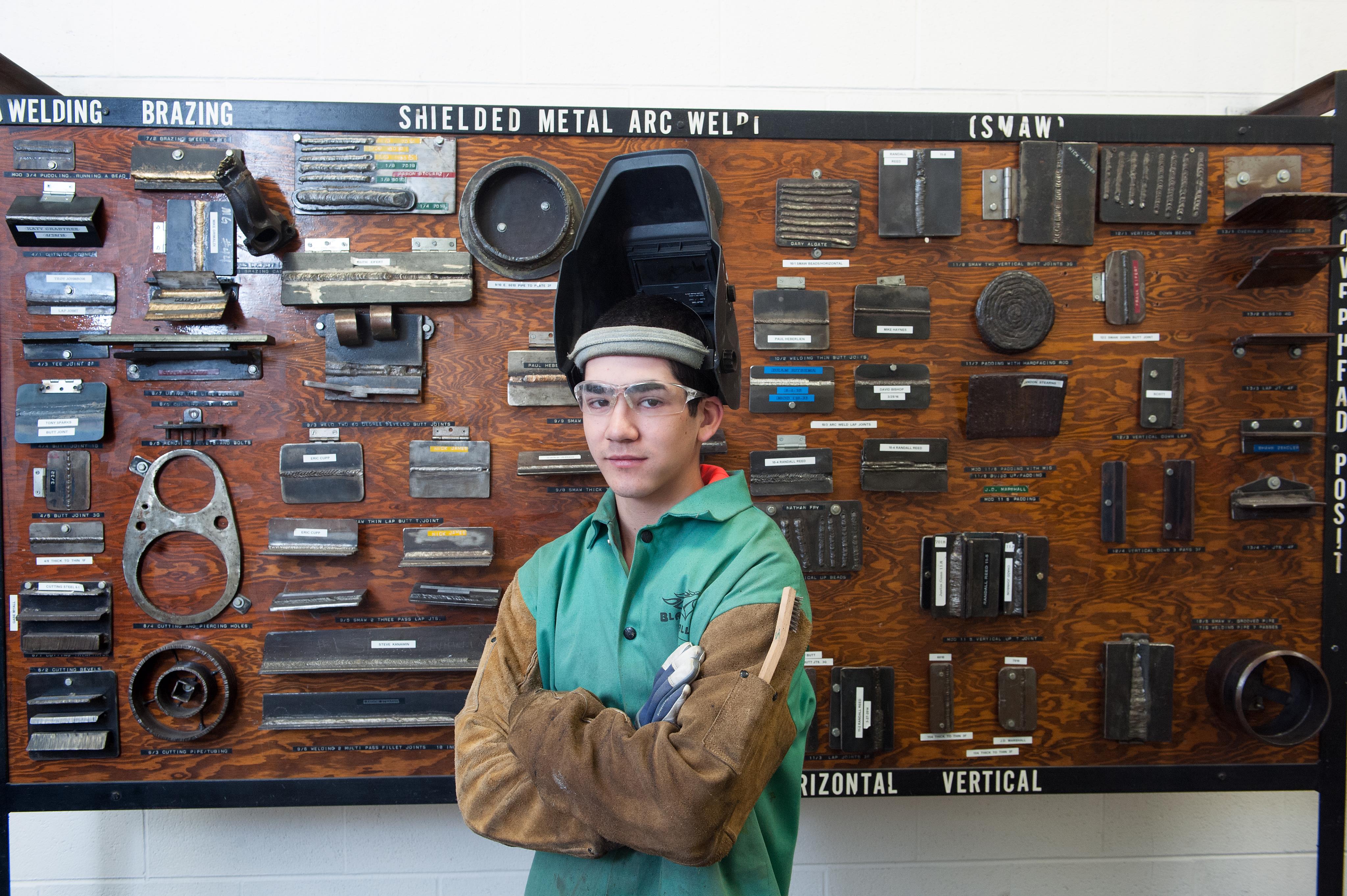 Student stands with arms folded in front of the Welding Wall of Fame board