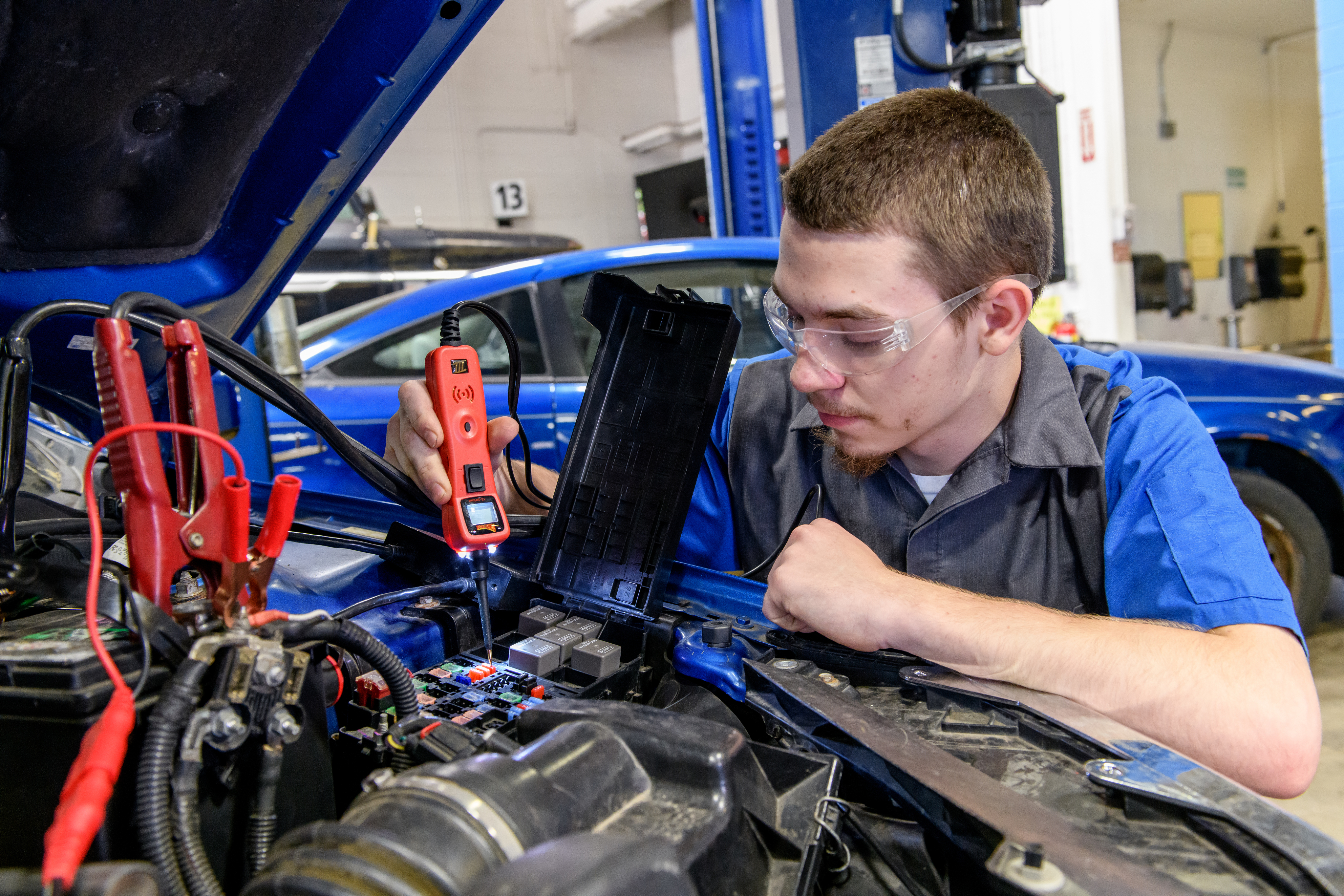 Automotive Technology student uses a tool to measure a car battery under the hood of a car.