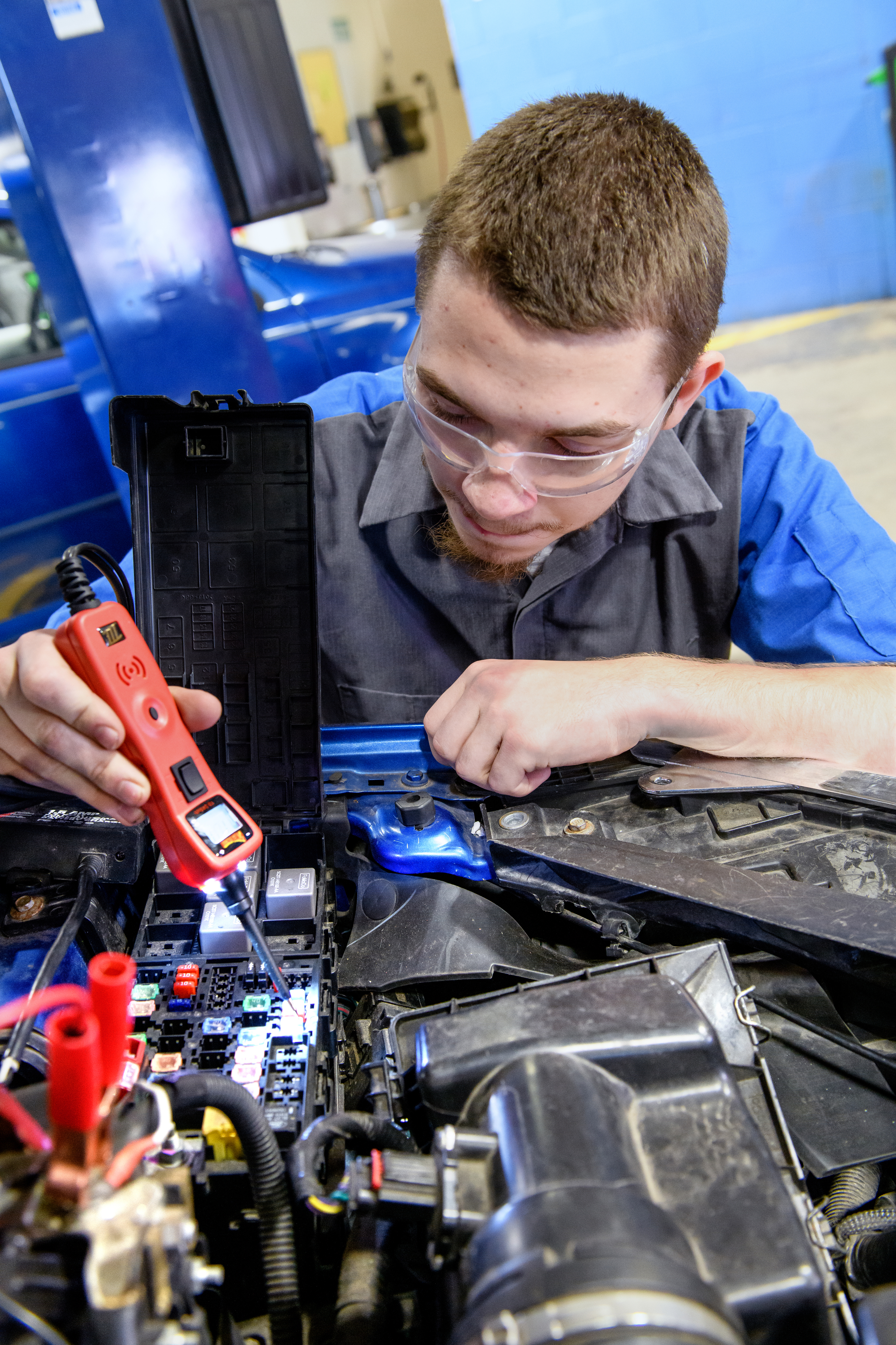 Male automotive technology student using an instrument to test a car battery under the hood of a blue car.