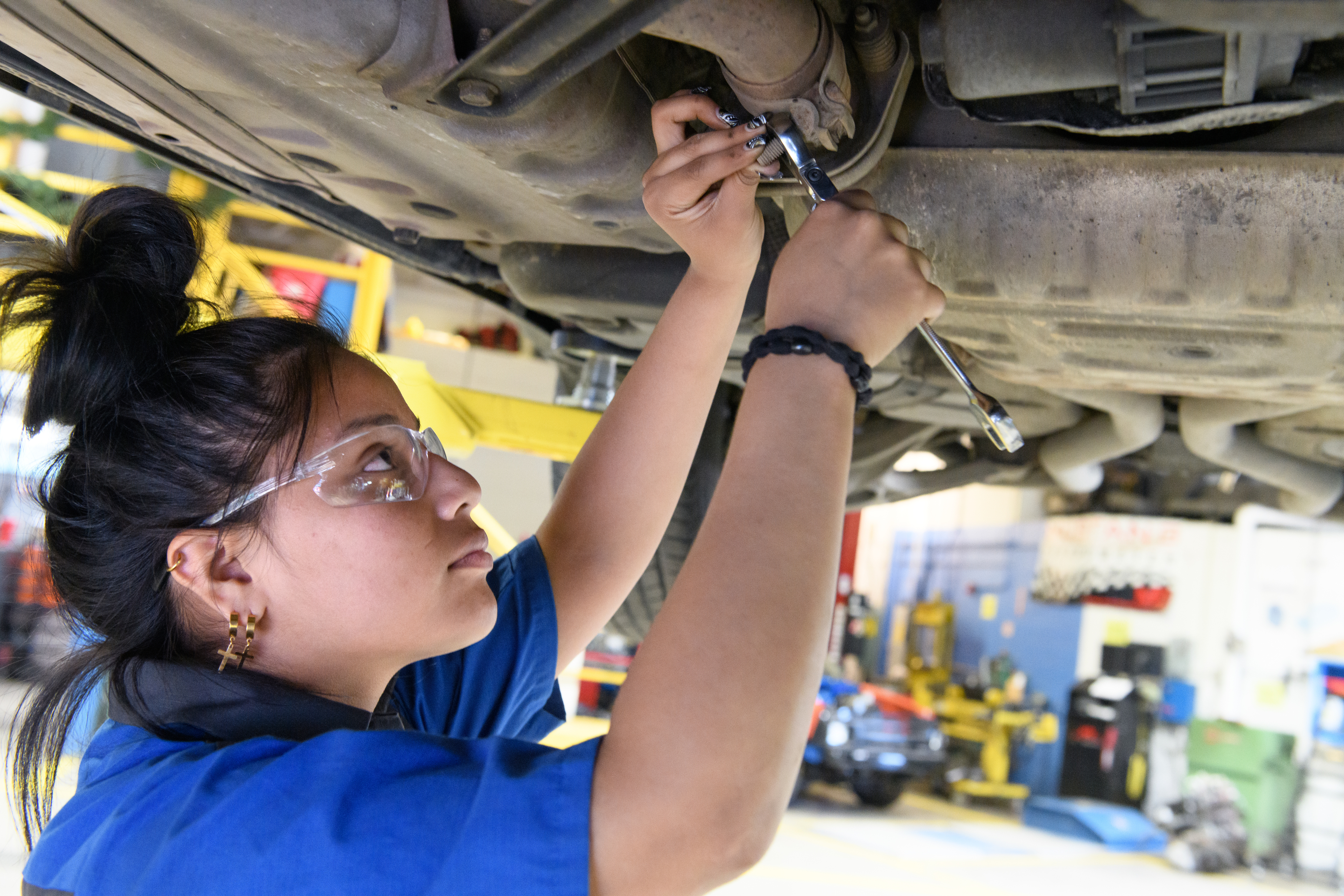 Female Automotive Technology student stands under a hoisted car, holding a wrench.