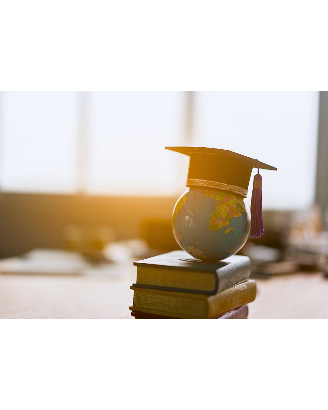 A graduation cap rests on a globe stacked on several books, with warm sunlight shining through a window in the background.